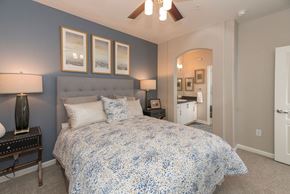 Sacramento apartment for rent - A serene bedroom here at Artisan 8282 featuring a plush upholstered bed with layered neutral bedding, matching nightstands with modern lamps, soft carpeting, and a calming accent wall, complemented by ceiling fan lighting and an arched opening leading to a bright en-suite bathroom.