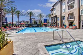 Corona apartment for rent - A resort-style outdoor pool here at Artisan at Main Street Metro featuring a large rectangular swimming pool with clear blue water, built-in steps and metal handrails, surrounded by red lounge chairs and umbrellas, tall palm trees, and modern apartment buildings under a bright blue sky with scattered clouds.