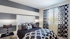 Corona apartment for rent - A stylish bedroom here at Artisan at Main Street Metro showcasing a tufted white headboard, bold striped accent wall in neutral tones, patterned black-and-white bedding, matching curtains framing a sunlit window, coordinated nightstands with modern lamps, and soft carpet flooring that creates a polished, inviting retreat.