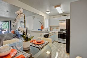 San Bernardino apartment for rent - A renovated kitchen and dining area here at Castlepark with white cabinets, light countertops, and a breakfast bar overlooking the space. The kitchen features a sink, stove, oven, microwave, refrigerator, and dishwasher, while the dining area includes a glass-top table set with place settings. Neutral finishes and overhead lighting create a clean, welcoming layout.