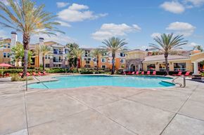 Moreno Valley apartment for rent - A resort-style swimming pool here at Fresco surrounded by tall palm trees, red lounge chairs, and wide concrete decking. The pool sits at the center of the community with Mediterranean-inspired apartment buildings and a clubhouse in the background, creating a bright, inviting outdoor space for relaxation and social gatherings.