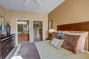 Moreno Valley apartment for rent - A spacious bedroom here at Fresco featuring plush carpet, a large bed with layered bedding, and a ceiling fan overhead. An open doorway leads to a walk-in closet with built-in shelving, while the ensuite bathroom with a dual-sink vanity is visible beyond, creating a comfortable and functional retreat.
