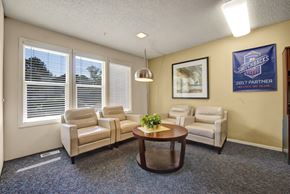 Colorado Springs apartment for rent - A cozy community lounge living room here at Austin Park featuring three cream leather armchairs arranged around a round wooden coffee table with greenery, soft carpet flooring, large windows with white blinds bringing in natural light, and warm accent walls with framed artwork and decor.