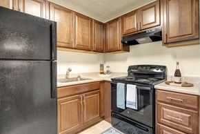 Colorado Springs apartment for rent - A warm, functional kitchen here at Austin Park featuring rich wood cabinetry, black appliances including a refrigerator and electric range, light countertops, a stainless sink, a vent hood, and neutral flooring, with a compact layout that offers efficient storage and a clean, practical space for everyday cooking and meal prep.