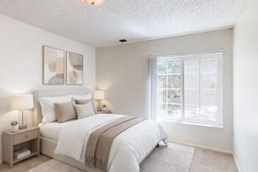 Colorado Springs apartment for rent - A bright, airy bedroom here at Austin Park featuring a softly upholstered bed with crisp white bedding, neutral accent pillows, matching bedside tables with modern lamps, abstract wall art above the headboard, plush carpet flooring, and a large window with vertical blinds that fills the space with natural light.