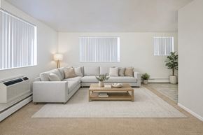 Colorado Springs apartment for rent - A bright modern living room here at Bristol Square featuring a spacious light gray sectional sofa with neutral throw pillows, a wooden coffee table on a soft area rug, vertical blinds allowing natural light, light-toned walls, and clean finishes that create an open, inviting atmosphere for everyday living and relaxing.