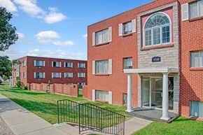 Colorado Springs apartment for rent - A classic brick apartment building exterior here at Bristol Square featuring a main entrance with white columns, an arched window above the doorway, multiple large windows with shutters, well-kept green lawns, a concrete walkway with railings, and additional residential buildings visible in the background under a bright blue sky.