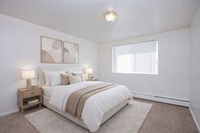 Colorado Springs apartment for rent - A calm and inviting bedroom here at Bristol Square featuring a queen-sized bed with crisp white bedding and a taupe throw blanket, matching wood nightstands with modern lamps, soft neutral wall art above the headboard, plush carpet flooring, and a wide window with vertical blinds that brings in gentle natural light.