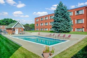 Colorado Springs apartment for rent - A sunny outdoor swimming pool here at Bristol Square featuring a rectangular pool surrounded by a concrete deck, red lounge chairs, green lawn landscaping, and a tall evergreen tree set against brick apartment buildings and a bright blue sky with scattered clouds.