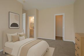 Colorado Springs apartment for rent - A cozy bedroom here at Camelback Pointe featuring soft neutral walls, plush carpet, and a light upholstered bed with layered white and beige bedding, complemented by simple wall art, a dresser for storage, and an en suite bathroom visible through the open doorway for added convenience.