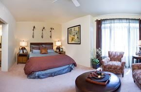 Colorado Springs apartment for rent - A cozy bedroom here at Cheyenne Creek Luxury showcasing a comfortable bed with layered bedding, matching nightstands with lamps, plush carpeting, and soft neutral walls, complemented by a sitting area with upholstered chairs, natural light from a large window with curtains, and tasteful wall art creating a relaxing retreat.