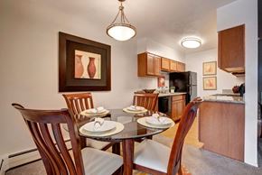 Colorado Springs apartment for rent - A welcoming dining area here at Copper Stone featuring a round glass-top table set for four with wooden chairs and upholstered seats, positioned beside an open kitchen with warm wood cabinetry, sleek countertops, modern appliances, pendant lighting, and framed wall art that creates a polished and comfortable apartment dining space.