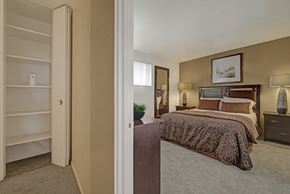 Colorado Springs apartment for rent - A warm bedroom here at Copper Stone showcasing a wood bed frame with patterned bedding, matching nightstands with lamps, soft carpet flooring, neutral wall tones, framed artwork above the headboard, a tall mirror, and an open shelving closet visible from the entry that adds functional storage and a comfortable, inviting layout.