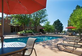 Colorado Springs apartment for rent - A bright outdoor pool area here at Eagle Ridge featuring a rectangular swimming pool with clear blue water, surrounding concrete deck, multiple lounge chairs arranged along the poolside, patio tables with red umbrellas, mature trees providing shade, and a clubhouse building visible in the background under a sunny blue sky.
