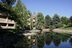 Colorado Springs apartment for rent - A peaceful landscaped pond here at Eagle Ridge reflecting surrounding trees and blue sky, with gently sloped grassy banks, natural stone edging, and mature evergreens alongside a brick apartment building with balconies, creating a serene outdoor setting that highlights the community’s greenery and scenic water feature.