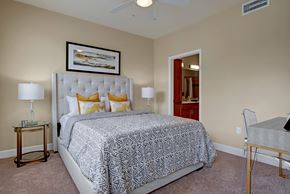 Colorado Springs apartment for rent - A serene bedroom here at Elements at Briargate featuring a plush upholstered bed with layered neutral bedding and gold accent pillows, matching bedside tables with modern lamps, soft carpeting, a ceiling fan overhead, and an open doorway leading to a private bathroom that enhances comfort and functionality.