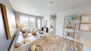 Colorado Springs apartment for rent - A contemporary living room here at Experience at Epicenter showcasing wood-style flooring, a light neutral sofa with accent pillows, a matching armchair, a round coffee table styled with decor, an abstract area rug, a floor lamp, framed wall art, open shelving with decorative accents, and large windows with a glass door that fill the space with natural light and offer balcony views.