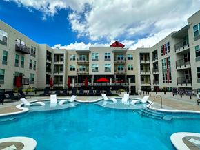 Colorado Springs apartment for rent - A resort-style outdoor pool here at Experience at Epicenter featuring a spacious swimming area with in-water lounge chairs, surrounding poolside seating, and red umbrellas for shade. Modern apartment buildings with balconies frame the courtyard, creating a vibrant community setting ideal for relaxing, socializing, and enjoying sunny days.