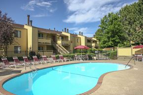 Colorado Springs apartment for rent - A sunny outdoor pool area here at Grand View featuring a curved swimming pool with clear blue water, brick-accented pool decking, red lounge chairs arranged along the fence, shaded patio tables with umbrellas, and landscaped greenery set in front of classic two-story apartment buildings under a bright blue sky.