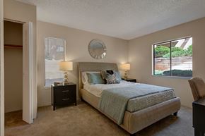 Colorado Springs apartment for rent - A bright and serene bedroom here at Grand View featuring a neatly made bed with soft blue and white bedding, a cushioned upholstered headboard, matching black nightstands with warm table lamps, plush carpeted flooring, a spacious open closet, and two windows that bring in natural light and views of greenery.