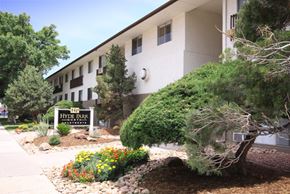 Colorado Springs apartment for rent - A landscaped community entrance here at Hyde Park North Apartments featuring a monument-style sign surrounded by colorful flowers, decorative rock beds, and mature trees. A multi-story apartment building with large windows sits behind the sign, creating a welcoming and well-maintained first impression of the community.