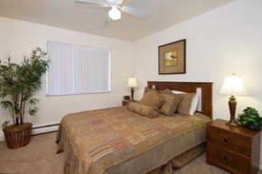 Colorado Springs apartment for rent - A comfortable bedroom here at Hyde Park North Apartments with beige carpet and light neutral walls, featuring a bed with patterned bedding, matching nightstands with lamps, and framed wall art above the headboard. A ceiling fan provides added comfort, while a window with vertical blinds and a potted plant create a calm, inviting atmosphere.
