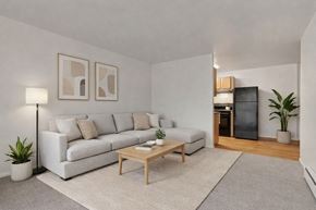 Colorado Springs apartment for rent - A bright, modern living room here at Hyde Park featuring a light gray sectional sofa with neutral accent pillows, a wooden coffee table centered on a soft area rug, simple abstract wall art, and floor and potted plants, with an open view into a compact kitchen with wood-look flooring and clean finishes.