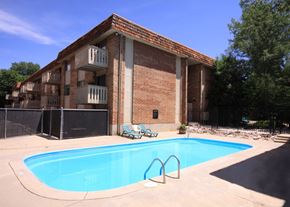 Colorado Springs apartment for rent - A refreshing outdoor pool area here at Hyde Park Apartments with a clear rectangular swimming pool surrounded by a concrete deck. Lounge chairs sit nearby for relaxing, while a brick apartment building with balconies and surrounding trees creates a private, inviting community atmosphere.