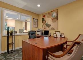 Colorado Springs apartment for rent - A welcoming leasing office here at Hyde Park Apartments with a wooden desk, guest chairs, and warm yellow walls. A large abstract art piece hangs above the desk, while a service window opens to an adjacent office area. Framed documents, soft lighting, and simple decor create a professional and inviting space for residents.