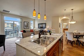 Colorado Springs apartment for rent - A bright, open kitchen and living area here at La Bella Vita featuring granite countertops with a double sink, warm wood cabinets, and pendant lighting over a spacious island with seating. The layout opens to a cozy living room with a sofa and balcony access, while wood-style flooring and a nearby dining area create an inviting, modern flow.