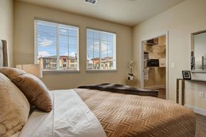 Colorado Springs apartment for rent - A serene primary bedroom here at La Bella Vita featuring a comfortable bed dressed in soft neutral bedding with layered pillows and a folded throw, positioned beside large windows with blinds that fill the room with natural light and views of the surrounding community. An open walk-in closet and simple accent furnishings complete the airy, inviting space.