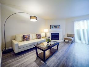 Colorado Springs apartment for rent - A bright living room here at Retreat at Austin Bluffs featuring a light-colored sofa with patterned accent pillows, a glass-top coffee table, and wide plank flooring. A curved floor lamp and table lamp add warm lighting, while a corner fireplace, sliding glass door with vertical blinds, and simple accent chair create a comfortable, open layout.