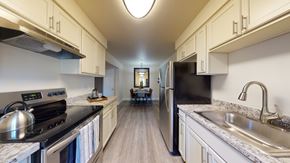 Colorado Springs apartment for rent - A bright galley-style kitchen here at Retreat at Austin Bluffs featuring white shaker-style cabinetry, speckled countertops, and wood-look flooring that extends toward a dining area. The space includes a refrigerator, range, sink, and modern lighting, creating a clean, functional layout with a warm, inviting feel.