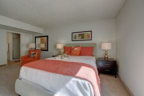 Colorado Springs apartment for rent - A cozy bedroom here at Retreat at Austin Bluffs featuring a comfortable upholstered bed with crisp white bedding, a coral quilt, and coordinating accent pillows. The space includes matching nightstands with modern lamps, an accent chair near the bathroom entry, soft carpet flooring, and warm natural light that creates a welcoming, relaxing atmosphere.