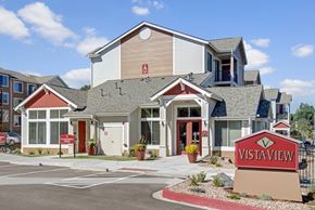 Colorado Springs apartment for rent - A welcoming exterior clubhouse building here at Vista View featuring peaked rooflines, neutral siding with red accent panels, large windows, and a clearly marked entrance, with landscaped planters, paved walkways, and a prominent Vista View monument sign set against a bright blue sky with scattered clouds.