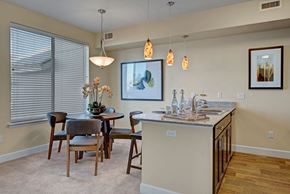 Colorado Springs apartment for rent - A bright open concept dining area here at Vista View featuring a round wooden dining table with four upholstered chairs, large window with blinds, and warm neutral walls, seamlessly connected to a kitchen island with stone-look countertops, modern pendant lighting, bar seating, and coordinated finishes that create a welcoming and functional space.
