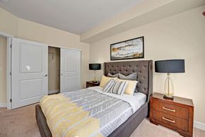 Colorado Springs apartment for rent - A cozy staged bedroom here at Vista View featuring a tufted upholstered bed with patterned neutral bedding, matching wood nightstands with modern lamps, soft carpet flooring, and sliding closet doors, complemented by warm wall tones and simple artwork that create a comfortable and inviting sleeping space.