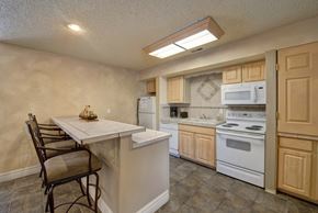 Colorado Springs apartment for rent - A cozy kitchen here at Windtree featuring light wood cabinetry, a peninsula-style counter with bar seating, white appliances, tiled flooring, and soft overhead lighting, creating a functional layout with ample counter space and an inviting area for casual dining and everyday cooking.
