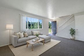 Colorado Springs apartment for rent - A bright modern living room here at Yorkshire Square featuring a light gray sectional sofa with neutral pillows, a simple wood coffee table on a soft area rug, and a large window bringing in natural light, with carpeted floors, an open stairway, and a welcoming entry that creates an airy, comfortable atmosphere.
