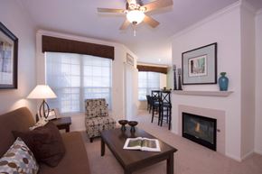 Colorado Springs apartment for rent - A bright, inviting living room here at Camelback Pointe featuring neutral carpeting, a cozy sofa with accent pillows, a patterned armchair, and a rectangular coffee table, with a fireplace, ceiling fan, framed artwork, and large windows that allow natural light to flow into the open dining area beyond.