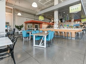 Minneapolis apartment for rent - A modern communal dining and bar area here at Blue and Lime featuring polished concrete floors, a long wood-accented bar with metal stools, and a central dining table surrounded by bright blue upholstered chairs. Industrial lighting, exposed ceilings, and greenery create an open, social atmosphere designed for gatherings and casual dining.