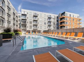 Minneapolis apartment for rent - A resort-style outdoor pool here at Blue and Lime featuring clear water surrounded by a spacious deck lined with orange lounge chairs and modern planters. Contemporary apartment buildings frame the courtyard, while railings, steps, and open sky create a bright, inviting setting designed for relaxation and social gathering.