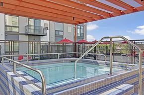 St. Louis Park apartment for rent - A modern outdoor spa here at 4800 Excelsior featuring a tiled hot tub with built-in steps and stainless handrails beneath a bold orange pergola. The fenced courtyard includes contemporary apartment façades, red patio umbrellas, and seating areas, creating a vibrant, inviting space for relaxation and socializing.