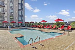 St. Louis Park apartment for rent - A resort-style outdoor pool here at 4800 Excelsior showcasing a rectangular swimming pool with clear blue water, stainless handrails, and a spacious concrete deck. Red lounge chairs and matching umbrellas line the poolside, with modern apartment buildings and treetop views creating an open, relaxing atmosphere.
