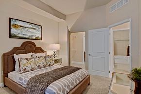 St. Louis Park apartment for rent - A serene bedroom here at 4800 Excelsior featuring a plush upholstered bed with patterned bedding, layered accent pillows, and a textured throw. Soft carpeting and neutral walls create a calming atmosphere, complemented by a bedside lamp, framed artwork, and a mirrored nightstand, with easy access to a walk-in closet and adjoining bathroom.