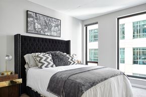 Minneapolis apartment for rent - A stylish bedroom here at 4Marq featuring a plush upholstered headboard with nailhead detailing, crisp white bedding layered with patterned accent pillows, and a soft gray throw. Large windows bring in natural light and city views, complemented by modern wall art, a sleek nightstand with a decorative lamp, and neutral carpeting.