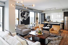 Minneapolis apartment for rent - A modern open-concept community lounge here at 4Marq showcasing a stylish seating area with a white sofa, patterned armchair, and leather accent chairs centered around a low coffee table. Floor-to-ceiling windows, exposed ductwork, contemporary lighting, and a sleek kitchen backdrop create a bright, urban atmosphere.