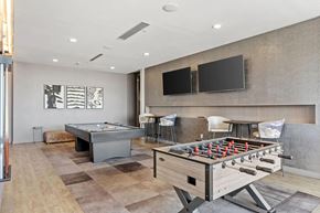 Minneapolis apartment for rent - A modern game lounge here at 4Marq featuring a foosball table in the foreground and a billiards table set on patterned flooring. Two wall-mounted flat-screen televisions sit above a sleek counter with bar-height seating, while neutral walls, framed abstract artwork, and recessed lighting create a relaxed and social atmosphere.