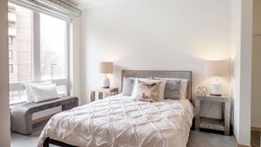 Minneapolis apartment for rent - A serene bedroom here at 7West featuring a neatly made bed with crisp white bedding and soft accent pillows, flanked by matching bedside tables with modern lamps. A large window with a built-in bench seat allows natural light to fill the space, while clean white walls, exposed ductwork, and polished concrete flooring create a calm, contemporary atmosphere.