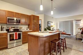 Sartell apartment for rent - A kitchen here at Club at Heritage featuring warm wood cabinetry, stainless steel appliances, and a spacious island with seating, opening into a bright living area with sliding glass doors and cozy furnishings.